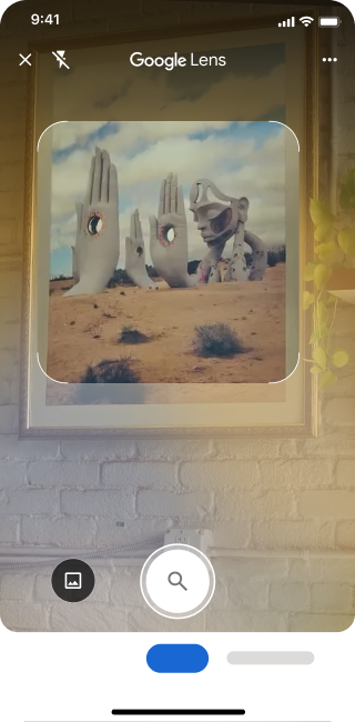 Sur un téléphone, l'application Google Lens est ouverte. Une notification indique à l'utilisateur qu'il peut appuyer sur le bouton de l'obturateur pour rechercher une image de grandes sculptures de mains en pierre dans un désert.